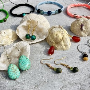 præsentation af håndlavede krystalsmykker på grå stenflade med hvide krystalgeoder. Bagerst, 4 kyttet armbånd med krystaller. Et i grøn, sort, lyseblå og rosa. 3 par creoløreringe med krystaller, 1 guld og 2 sølv. 3 par øreringe, 2 guld og 1 sølv.