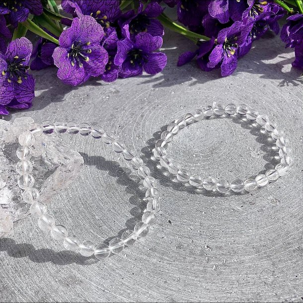 To gennemsigtige bjergkrystal armb�nd med 6mm perler, der ligger smukt ved siden af hinanden p� en gr� overflade.