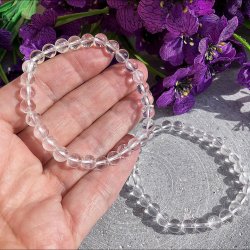 N�rbillede af et krystalklart bjergkrystal armb�nd i en h�nd med lilla blomster i baggrunden.