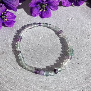 Fint og spinkelt regnbue fluorit armb�nd med 4 mm transparente krystalperler liggende p� en gr� baggrund pyntet med lilla blomster i toppen.