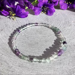 Rundt regnbue fluorit krystalarmb�nd med 4 mm perler fotograferet p� en gr� overflade med dekorative lilla blomster.