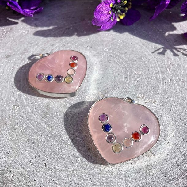 Hjerteformede rosakvarts vedh�ng med de syv chakra sten fotograferet p� en gr� baggrund med lilla blomster.