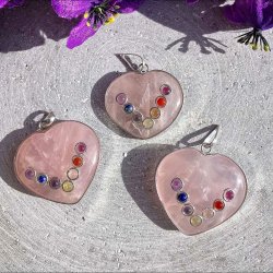 Tre hjerteformede rosakvarts vedh�ng med de syv chakra sten samlet p� en gr� rustik overflade.